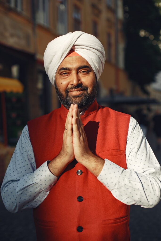 Smiling man in traditional attire and turban with a greeting pose outdoors.